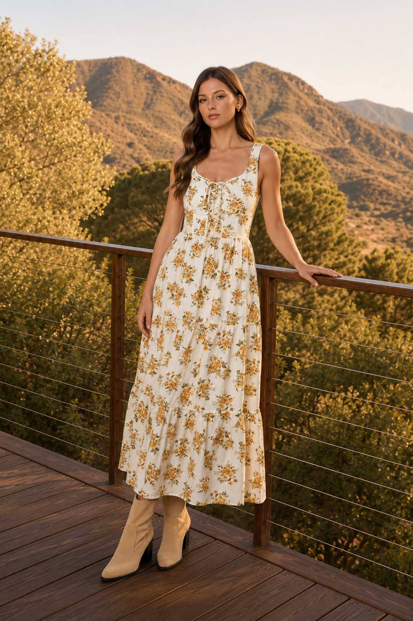 Woman wearing a cream floral sleeveless midi dress with a lace-up corset bodice, tiered skirt, and ankle boots, standing outdoors with mountain views in the background.