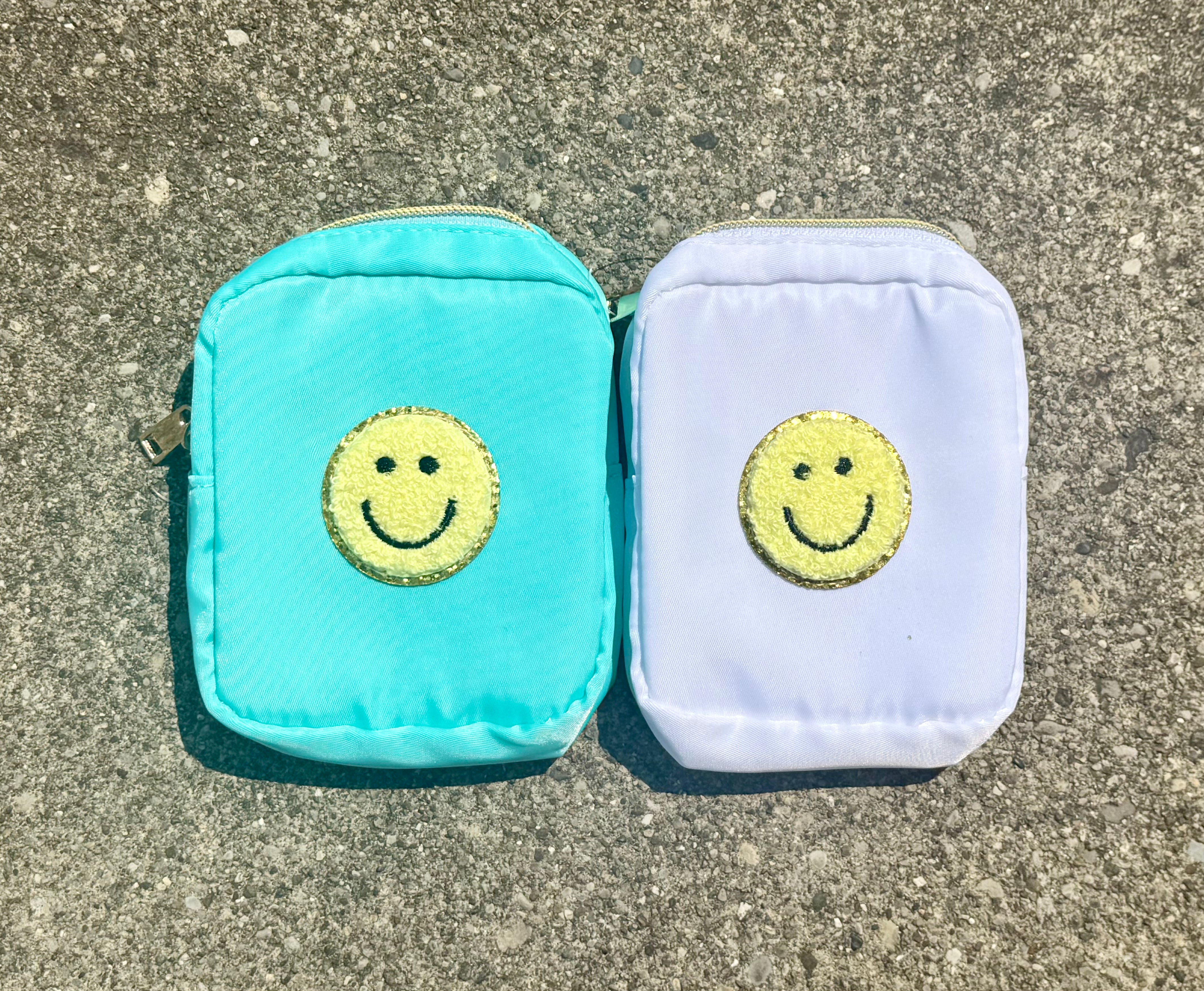 Two small zippered pouches with embroidered smiley faces, one in turquoise and one in pale lavender, placed side by side on a gray concrete surface.