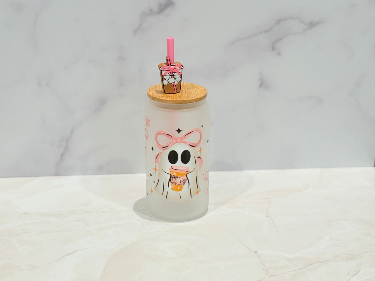 A frosted glass tumbler with a cute cartoon skeleton character holding a drink, topped with a bamboo lid and a small pink drink accessory, set on a light marble surface against a marble background.