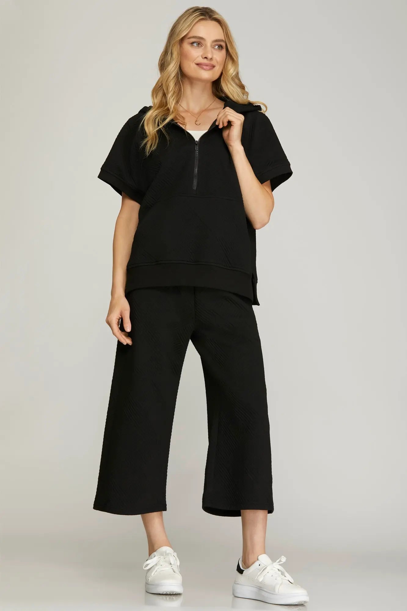 Women’s black quilted half-zip hoodie lounge set featuring a short-sleeve hooded top, cropped wide-leg pants, and white sneakers. Comfortable casual outfit perfect for travel, errands, and everyday wear.