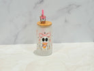 A frosted glass tumbler with a cute cartoon skeleton character holding a drink, topped with a bamboo lid and a small pink drink accessory, set on a light marble surface against a marble background.