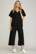 Women’s black quilted half-zip hoodie lounge set featuring a short-sleeve hooded top, cropped wide-leg pants, and white sneakers. Comfortable casual outfit perfect for travel, errands, and everyday wear.