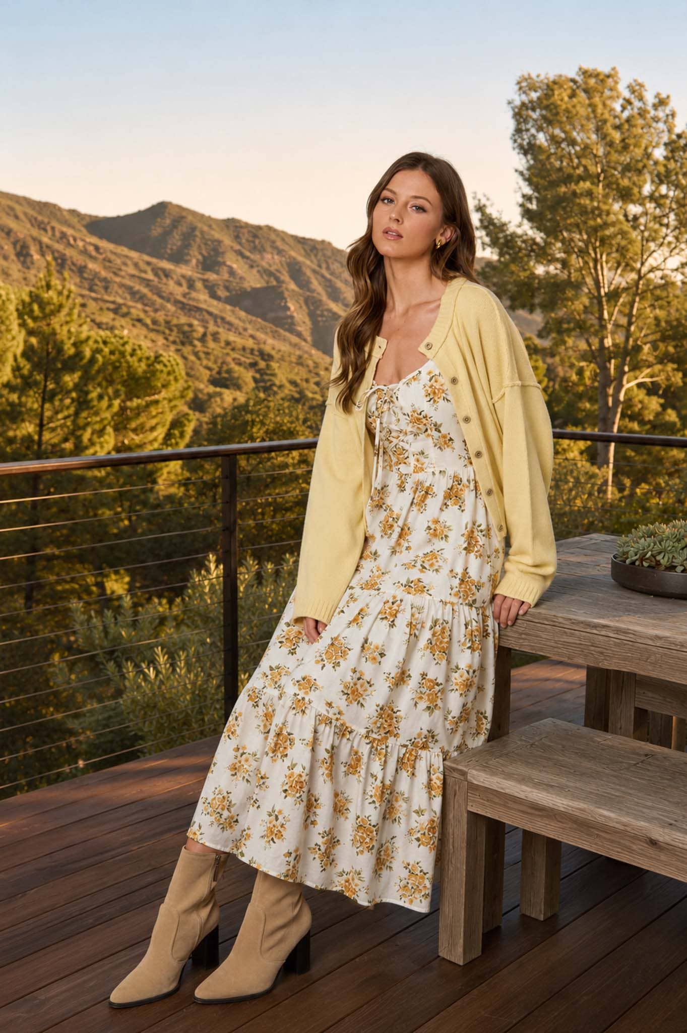 Woman in a floral dress and yellow cardigan standing on a wooden deck with mountains in the background